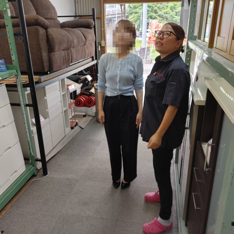 KKB取材で店内のソファを見上げるレポーターとリサイクルショップ八光スタッフ（鹿児島市田上）